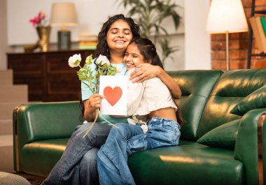 Hintli Asyalı genç anne, küçük kızına kanepede sarılıp, kalpten bir Anneler Günü 'nü kutlarken, kızının yaptığı çiçekler ve hediyelere bakarken aile bağlarını sergiliyor, sevgisini sergiliyor.