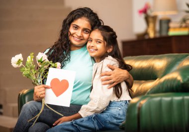 Hintli Asyalı genç anne, küçük kızına kanepede sarılıp, kalpten bir Anneler Günü 'nü kutlarken, kızının yaptığı çiçekler ve hediyelere bakarken aile bağlarını sergiliyor, sevgisini sergiliyor.