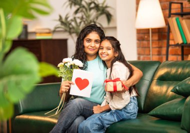 Hintli Asyalı genç anne, küçük kızına kanepede sarılıp, kalpten bir Anneler Günü 'nü kutlarken, kızının yaptığı çiçekler ve hediyelere bakarken aile bağlarını sergiliyor, sevgisini sergiliyor.