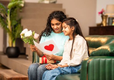 Hintli Asyalı genç anne, küçük kızına kanepede sarılıp, kalpten bir Anneler Günü 'nü kutlarken, kızının yaptığı çiçekler ve hediyelere bakarken aile bağlarını sergiliyor, sevgisini sergiliyor.