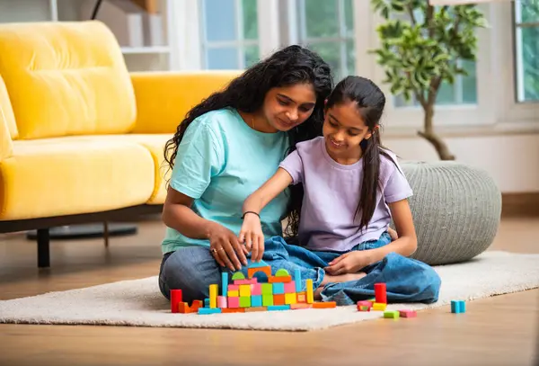 Hint Asyalı genç anne ve küçük kızı renkli ahşap oyuncaklar kullanarak ev inşa ederken modern müsrif bir evin halılı zemininde birlikte oturuyorlar ve kapalı alanda kaliteli zaman geçiriyorlar.