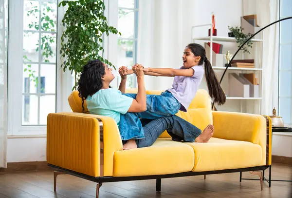 Hintli Asyalı genç anne küçük kızıyla modern oturma odasındaki kanepede neşeyle tahterevalli gibi oynuyor, eğlenceli bağlılık anları, mutluluk, kahkaha ve kapalı alanda kaliteli zaman geçirmenin tadını çıkarıyor.