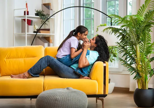 Hintli Asyalı genç anne küçük kızıyla modern oturma odasındaki kanepede neşeyle tahterevalli gibi oynuyor, eğlenceli bağlılık anları, mutluluk, kahkaha ve kapalı alanda kaliteli zaman geçirmenin tadını çıkarıyor.