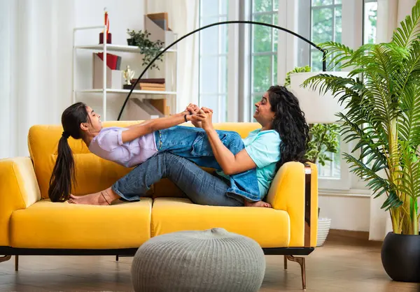 Hintli Asyalı genç anne küçük kızıyla modern oturma odasındaki kanepede neşeyle tahterevalli gibi oynuyor, eğlenceli bağlılık anları, mutluluk, kahkaha ve kapalı alanda kaliteli zaman geçirmenin tadını çıkarıyor.