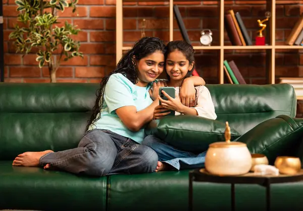 Hintli Asyalı genç anne kız evdeki rahat modern koltukta oturuyor, akıllı telefonu online alışveriş için kullanıyor, interneti tarıyor, sosyal medyayı karıştırıyor, video görüşmeleri yapıyor, dijital yaşam tarzının tadını çıkarıyor.