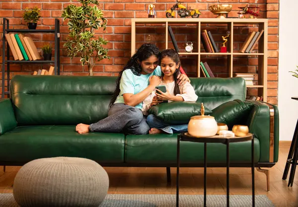 Hintli Asyalı genç anne kız evdeki rahat modern koltukta oturuyor, akıllı telefonu online alışveriş için kullanıyor, interneti tarıyor, sosyal medyayı karıştırıyor, video görüşmeleri yapıyor, dijital yaşam tarzının tadını çıkarıyor.