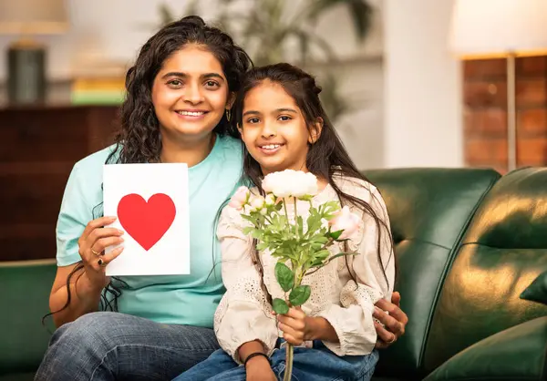 Hintli Asyalı genç anne, küçük kızına kanepede sarılıp, kalpten bir Anneler Günü 'nü kutlarken, kızının yaptığı çiçekler ve hediyelere bakarken aile bağlarını sergiliyor, sevgisini sergiliyor.