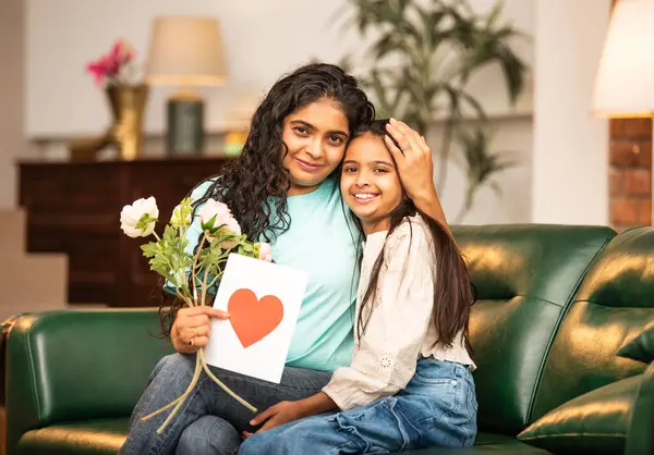 Hintli Asyalı genç anne, küçük kızına kanepede sarılıp, kalpten bir Anneler Günü 'nü kutlarken, kızının yaptığı çiçekler ve hediyelere bakarken aile bağlarını sergiliyor, sevgisini sergiliyor.