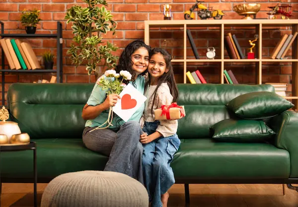 Hintli Asyalı genç anne, küçük kızına kanepede sarılıp, kalpten bir Anneler Günü 'nü kutlarken, kızının yaptığı çiçekler ve hediyelere bakarken aile bağlarını sergiliyor, sevgisini sergiliyor.