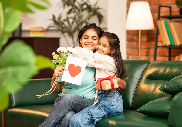 Hintli Asyalı genç anne, küçük kızına kanepede sarılıp, kalpten bir Anneler Günü 'nü kutlarken, kızının yaptığı çiçekler ve hediyelere bakarken aile bağlarını sergiliyor, sevgisini sergiliyor.