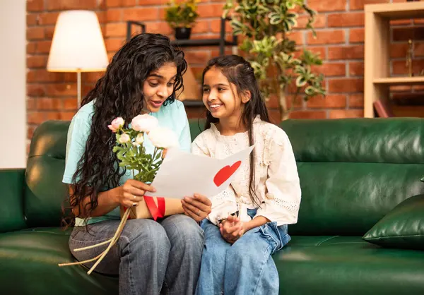 Hintli Asyalı genç anne, küçük kızına kanepede sarılıp, kalpten bir Anneler Günü 'nü kutlarken, kızının yaptığı çiçekler ve hediyelere bakarken aile bağlarını sergiliyor, sevgisini sergiliyor.