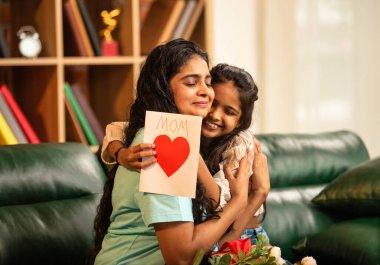 Hintli Asyalı genç anne, küçük kızına kanepede sarılıp, kalpten bir Anneler Günü 'nü kutlarken, kızının yaptığı çiçekler ve hediyelere bakarken aile bağlarını sergiliyor, sevgisini sergiliyor.