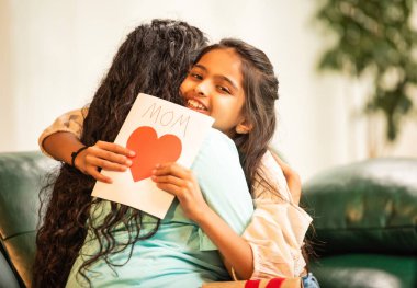 Hintli Asyalı genç anne, küçük kızına kanepede sarılıp, kalpten bir Anneler Günü 'nü kutlarken, kızının yaptığı çiçekler ve hediyelere bakarken aile bağlarını sergiliyor, sevgisini sergiliyor.