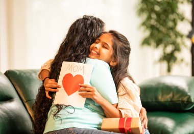 Hintli Asyalı genç anne, küçük kızına kanepede sarılıp, kalpten bir Anneler Günü 'nü kutlarken, kızının yaptığı çiçekler ve hediyelere bakarken aile bağlarını sergiliyor, sevgisini sergiliyor.