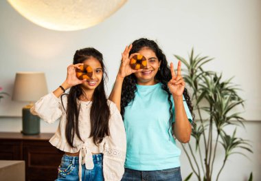 Hintli Asyalı genç bir anne ve şirin küçük bir kız kanepede oturuyor modern bir evde, neşeyle yuvarlak bir çörek tutuyor, delikten bakıyor, gülümsüyor ve iyi bir el hareketi yapıyor.