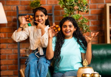 Hintli Asyalı genç bir anne ve şirin küçük bir kız kanepede oturuyor modern bir evde, neşeyle yuvarlak bir çörek tutuyor, delikten bakıyor, gülümsüyor ve iyi bir el hareketi yapıyor.