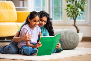 Hintli Asyalı genç anne ve şirin küçük kız şık, modern bir oturma odasında birlikte oturmuş, ilgi ve neşe dolu bir kitap okuyor, eğitici bir bağ kurma zamanının keyfini çıkarıyor, aşkı besliyor.