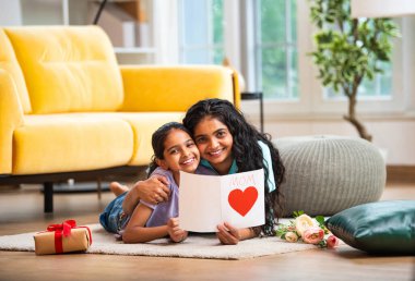 Hintli Asyalı genç anne ve küçük kızı modern şık bir evde, halının üzerinde uzanmış neşeli bir Anneler Günü kartı okuyorlar. Aile bağlarını paylaşıyorlar.