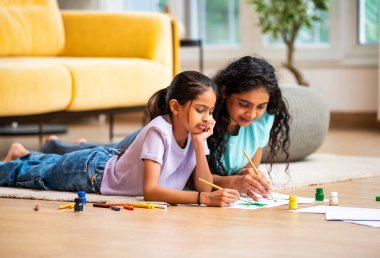 Hintli Asyalı genç anne ve küçük kızı modern bir evin zemininde uzanmış, kağıt üzerinde fırça ve su renklerini kullanarak sanat, çizim ve resim faaliyetleriyle mutlu mesut ilgileniyorlar.