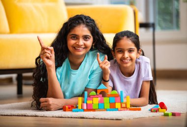Hint Asyalı genç anne ve küçük kızı renkli ahşap oyuncaklar kullanarak ev inşa ederken modern müsrif bir evin halılı zemininde birlikte oturuyorlar ve kapalı alanda kaliteli zaman geçiriyorlar.