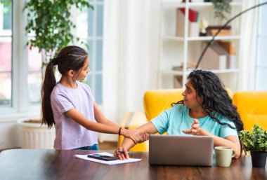 Hintli Asyalı genç anne, modern evdeki dizüstü bilgisayarında çalışırken, sevimli küçük kızı dikkat çekmek için elini çekerek, iş-yaşam dengesi mücadelesi sergiliyor.