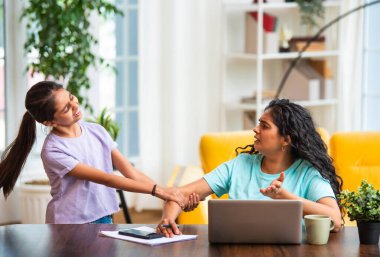 Hintli Asyalı genç anne, modern evdeki dizüstü bilgisayarında çalışırken, sevimli küçük kızı dikkat çekmek için elini çekerek, iş-yaşam dengesi mücadelesi sergiliyor.