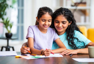 Hintli Asyalı küçük kız, modern bir evde masasında otururken pastel boya kalemleriyle resim çizmeye odaklanmış. Genç annesi de onun yaratıcılığını, sanatını ve adanmışlığını takdir ediyor.