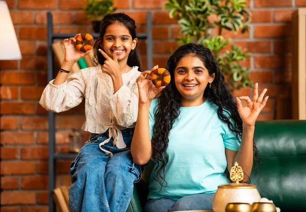 Hintli Asyalı genç bir anne ve şirin küçük bir kız kanepede oturuyor modern bir evde, neşeyle yuvarlak bir çörek tutuyor, delikten bakıyor, gülümsüyor ve iyi bir el hareketi yapıyor.