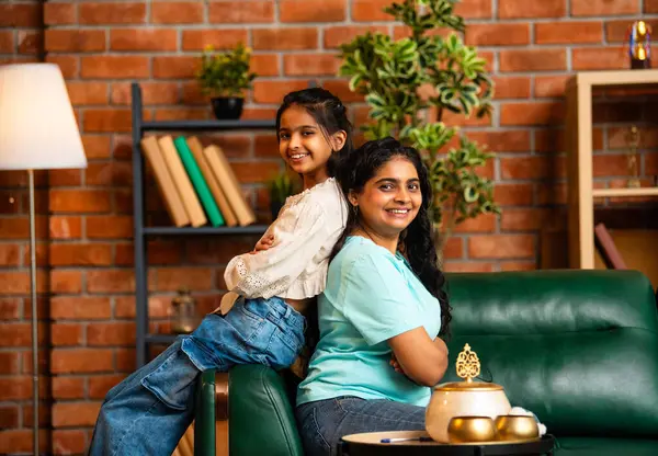 Hintli şirin küçük kız ve genç anne, rahat bir kanepede sırtüstü oturuyor, şakacı bir şekilde birbirlerini sevgiyle itiyorlar, eğlenceli, neşeli ve sevgi dolu bir anı paylaşıyorlar.