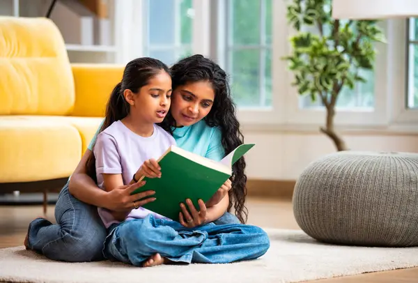 Hintli Asyalı genç anne ve şirin küçük kız şık, modern bir oturma odasında birlikte oturmuş, ilgi ve neşe dolu bir kitap okuyor, eğitici bir bağ kurma zamanının keyfini çıkarıyor, aşkı besliyor.