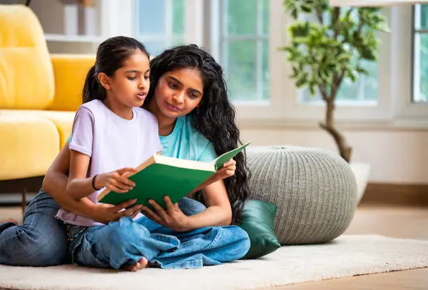 Hintli Asyalı genç anne ve şirin küçük kız şık, modern bir oturma odasında birlikte oturmuş, ilgi ve neşe dolu bir kitap okuyor, eğitici bir bağ kurma zamanının keyfini çıkarıyor, aşkı besliyor.