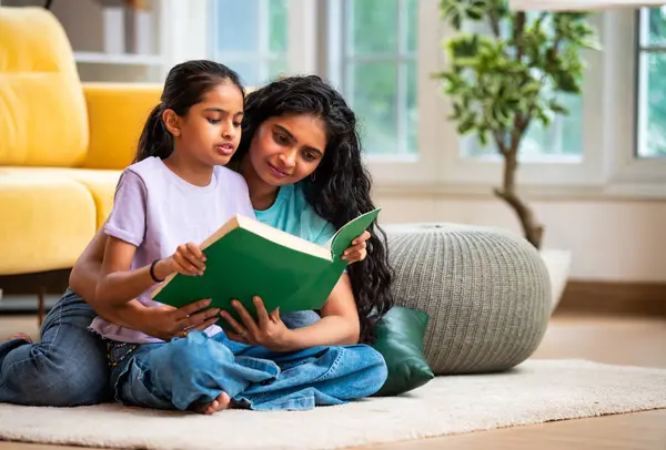 Hintli Asyalı genç anne ve şirin küçük kız şık, modern bir oturma odasında birlikte oturmuş, ilgi ve neşe dolu bir kitap okuyor, eğitici bir bağ kurma zamanının keyfini çıkarıyor, aşkı besliyor.