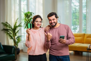 Hintli Asyalı genç çift, online alışveriş veya ödül kazandıktan sonra kredi veya banka kartı akıllı telefonunu tutarken modern oturma odasında neşeli ifadeler sergilerken başarıyı kutluyorlar.