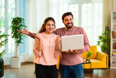 Hintli Asyalı çekici genç çift kablosuz bilgisayar kullanıyor, internette geziniyor ve online başarıyı kutluyor ya da neşeli ifadeler ve neşeli ruh haliyle oturma odasında duruyor.