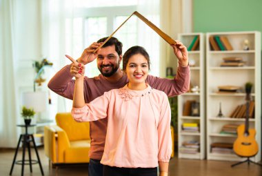 Hintli Asyalı koca, mutlu eşinin kafasının üstünde sembolik bir karton çatı yaratıyor. Yeni ev konseptinde yeni ev konseptini temsil ederken aynı zamanda oturma odasında bir arada duruyor.