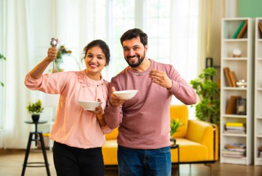 Hintli Asyalı genç çift kaselerden birlikte lezzetli yemekler yiyorlar. Kaşıkla, sevinçle, tatminle, yemek saatinde kaynaşarak modern oturma odasında duruyorlar.