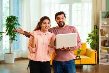 Hintli Asyalı çekici genç çift kablosuz bilgisayar kullanıyor, internette geziniyor ve online başarıyı kutluyor ya da neşeli ifadeler ve neşeli ruh haliyle oturma odasında duruyor.