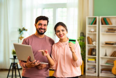 Hintli Asyalı genç çift ellerinde dizüstü bilgisayar, kredi kartı ya da banka kartı tutarak evde online ödeme başarısını kutluyor, neşeyle gülümsüyor ve modern oturma odasının iç kısmında olumlu ifadeler sergiliyorlar.