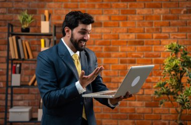 Hintli iş adamı çevrimiçi video görüşmesi sırasında dizüstü bilgisayarını kullanarak başarıyı kutlarken aynı zamanda güvenli bir şekilde duruyor ve terracotta tuğladan duvarlı rahat modern ofiste mutluluk ve neşe ifade ediyor.