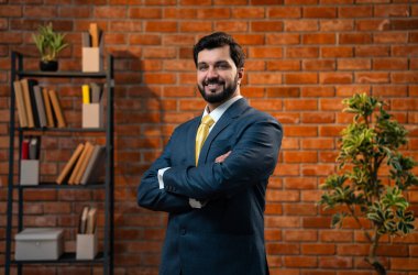 Mutlu Hintli genç işadamı veya CEO 'su sakallı, kollarını kavuşturmuş, terracotta tuğla duvarlı, rahat modern bir ofiste gururlu ve başarılı görünüyor.