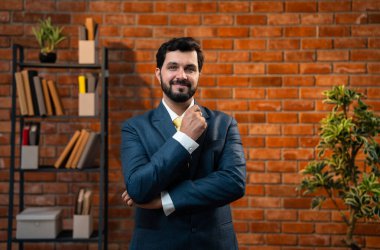 Mutlu Hintli genç işadamı veya CEO 'su sakallı, kollarını kavuşturmuş, terracotta tuğla duvarlı, rahat modern bir ofiste gururlu ve başarılı görünüyor.