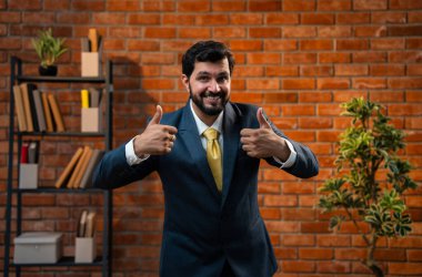 Başparmak, yumruk ya da zafer işareti, resmi takım elbiseli ve sakallı Hintli mutlu Asyalı işadamı ya da CEO terracotta tuğladan duvarlı samimi modern ofiste durarak başarıyı kutluyor.