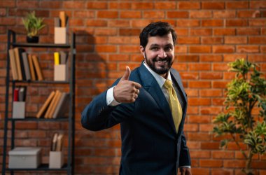 Başparmak, yumruk ya da zafer işareti, resmi takım elbiseli ve sakallı Hintli mutlu Asyalı işadamı ya da CEO terracotta tuğladan duvarlı samimi modern ofiste durarak başarıyı kutluyor.