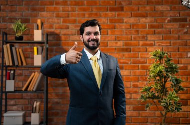 Başparmak, yumruk ya da zafer işareti, resmi takım elbiseli ve sakallı Hintli mutlu Asyalı işadamı ya da CEO terracotta tuğladan duvarlı samimi modern ofiste durarak başarıyı kutluyor.