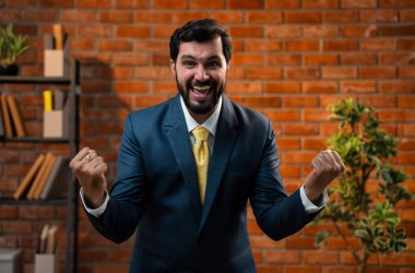 Başparmak, yumruk ya da zafer işareti, resmi takım elbiseli ve sakallı Hintli mutlu Asyalı işadamı ya da CEO terracotta tuğladan duvarlı samimi modern ofiste durarak başarıyı kutluyor.