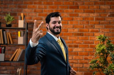 Başparmak, yumruk ya da zafer işareti, resmi takım elbiseli ve sakallı Hintli mutlu Asyalı işadamı ya da CEO terracotta tuğladan duvarlı samimi modern ofiste durarak başarıyı kutluyor.