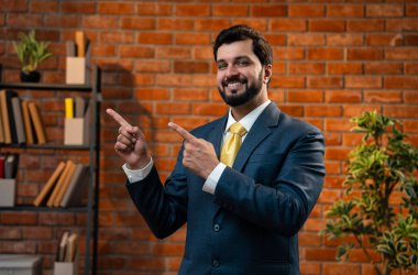 Mutlu Hintli Asyalı genç işadamı ya da CEO 'su sakallı, güvenilir bir şekilde gülümseyen ve yan tarafları işaret eden rahat bir ofiste dururken bir şeyleri destekleyen, reklamını yapan ya da onaylayan.