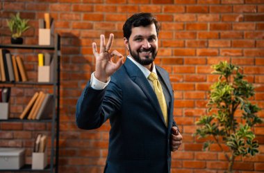 El işaretini gösteren mutlu Hintli genç işadamı veya CEO 'su sakallı resmi kurumsal kıyafetle rahat modern ofiste sıcak terracotta tuğlalı duvarlı kameraya bakıyor.