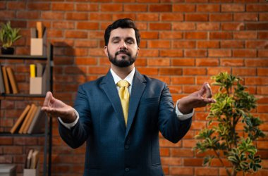 namaste pozunda katlanan eller, Hint Asyalı genç işadamı veya CEO kurumsal giysiler içinde sakin bir şekilde meditasyon yapıyor modern ofiste terracotta tuğlalı duvara karşı stres atağı ve farkındalık için