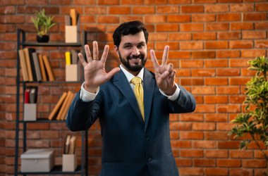 Sayıları gösterirken kendine güvenen parmakları kullanan, resmi kurumsal giyinmiş Hintli Asyalı sakallı ceo işadamı parmaklarını sayarken tuğla duvara karşı samimi bir ofiste duruyor.